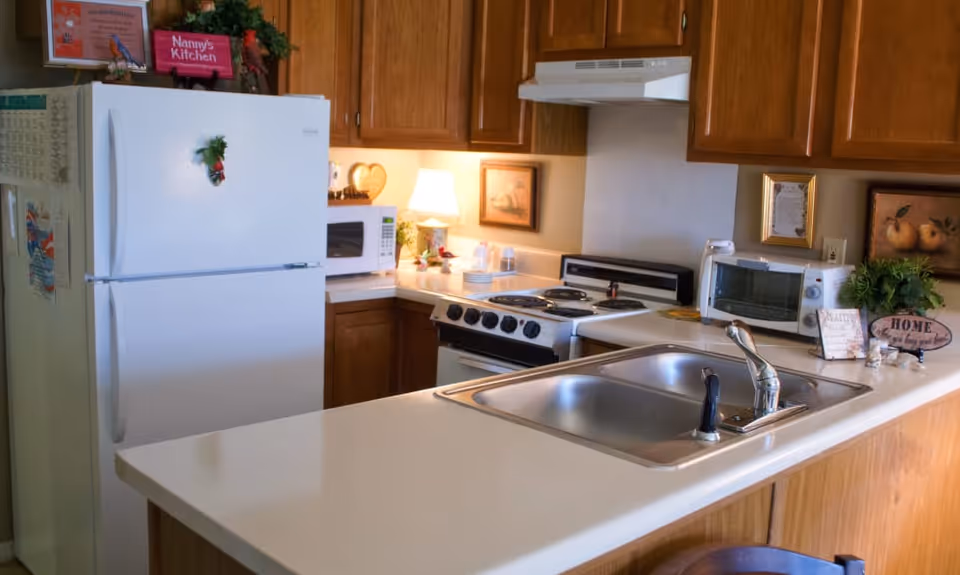A cozy kitchen with wooden cabinets, a white refrigerator decorated with a small wreath, a microwave, an electric stove with four burners, a toaster oven, and a double sink on a white countertop. The kitchen is decorated with small framed pictures, a lamp, and a small plant with a sign that says 'HOME'.
