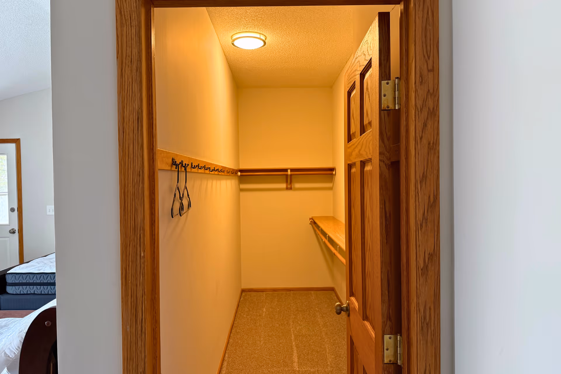 View into a small walk-in closet with beige walls and carpeted floor. The closet has wooden trim and a wooden door that is partially open. Inside, there are wooden shelves and a row of hooks on the left wall with a few hangers. Part of a bedroom with a bed and a door leading outside is visible to the left of the closet.