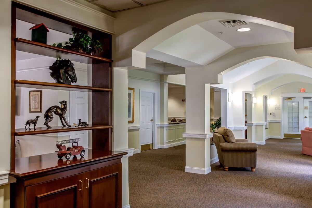 Interior view of a senior living facility hallway with beige walls and carpeted floors. There is a wooden shelving unit on the left displaying decorative items including horse and dog figurines and a small red toy fire truck. The hallway features white columns and arches, with a brown upholstered armchair and a pink sofa visible in the background. Wall sconces provide lighting along the corridor.