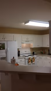 A clean and well-lit kitchen with white cabinets, a white refrigerator, a white stove with a range hood, and a countertop with a few kitchen items including a toaster and utensils. There is a fluorescent ceiling light and a breakfast bar in the foreground.