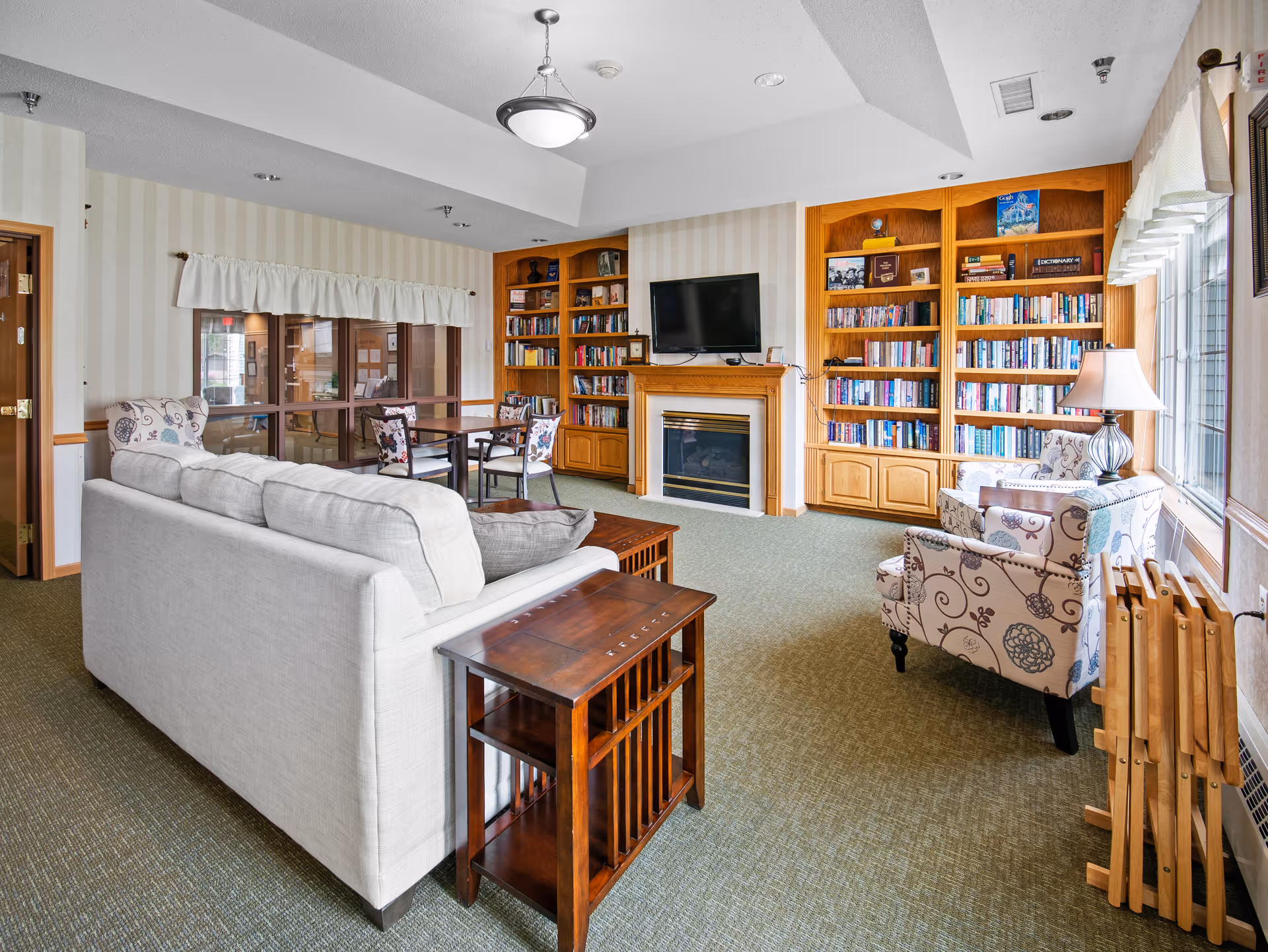 A cozy living room area in a senior living facility featuring a white sofa, two patterned armchairs, a wooden side table, and a fireplace with a mounted flat-screen TV above it. There are built-in wooden bookshelves filled with books on either side of the fireplace. A window with white curtains allows natural light to brighten the room. A small dining table with chairs is visible in the background.