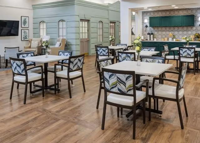 Communal dining room with square tables and patterned chairs on wood flooring, with a kitchenette and seating area in the background.