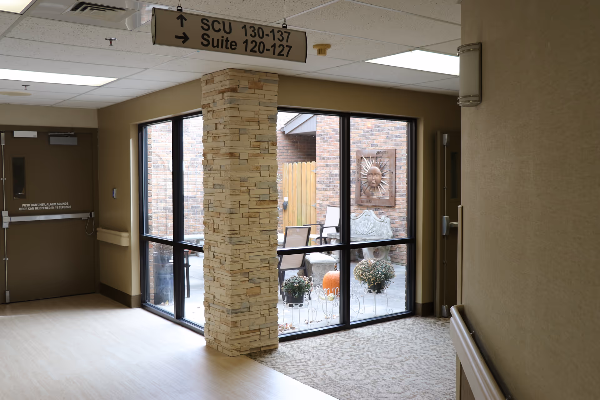 A senior living facility hallway with a stone column and overhead directional sign beside large windows looking into a small courtyard with a bench, sun wall art, potted plants and a pumpkin.