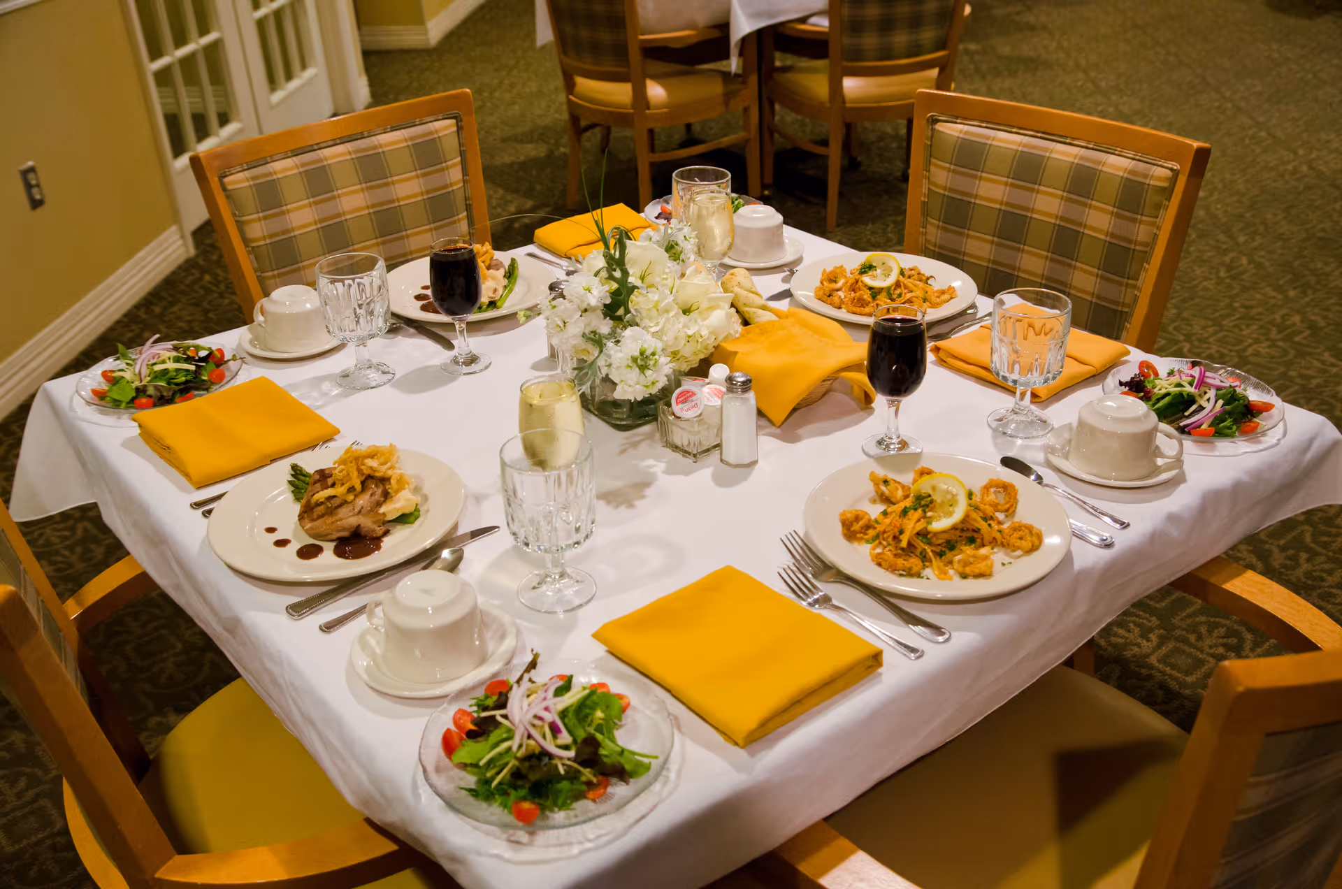 A dining table set for four with plated meals, salads, glasses, yellow napkins, and a floral centerpiece in a dining room.