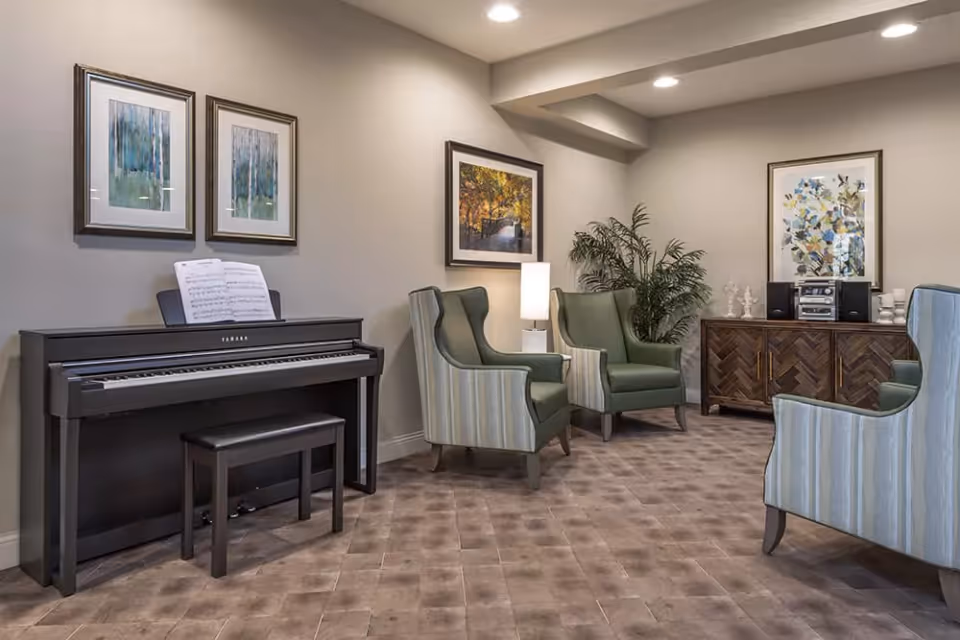 A cozy living room area with a black Yamaha piano and bench on the left, three green and striped armchairs arranged around a wooden sideboard with a stereo system, decorative items, and a potted plant. The walls are decorated with framed artwork and the floor has a patterned carpet.