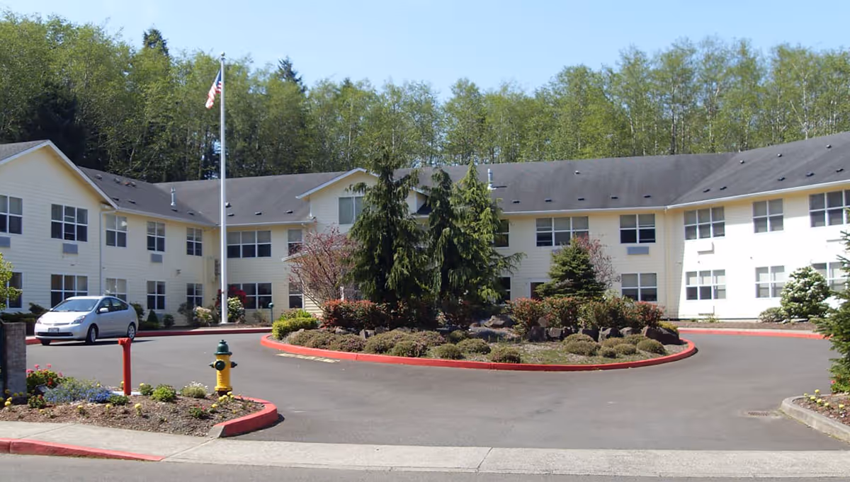 Exterior view of a two-story assisted living community building with beige siding and multiple windows. There is a circular driveway with landscaped greenery and trees in the center, an American flag on a flagpole, a fire hydrant, and a parked silver car. Trees and forested area are visible in the background under a clear blue sky.