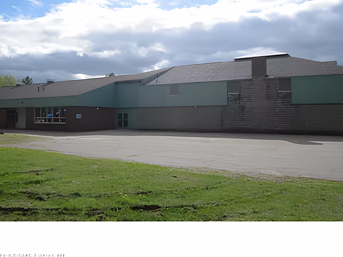 Exterior view of a large building with a sloped roof, green and brown walls, a paved area in front, and a grassy lawn in the foreground under a partly cloudy sky.