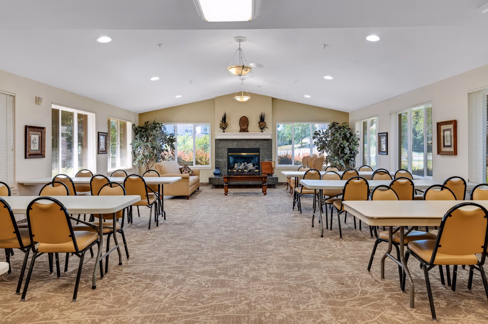 A spacious, well-lit common room with multiple tables and chairs arranged in rows. The room features large windows on both sides letting in natural light, two beige sofas near a central fireplace with decorative items on the mantel, and potted plants flanking the fireplace. The ceiling has recessed lighting and two hanging light fixtures.