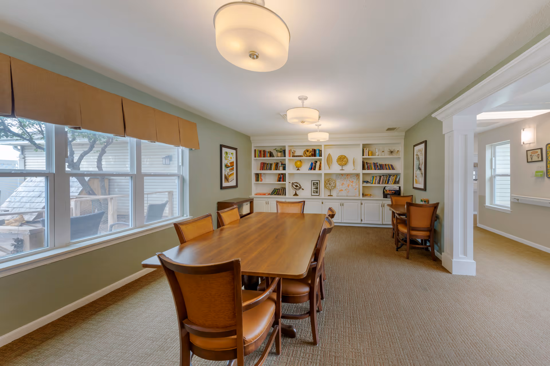 A well-lit room with a long wooden table surrounded by eight chairs with brown leather seats. The room has a large window with a brown valance, beige carpet, and light green walls. At the far end, there is a white built-in bookshelf filled with books and decorative items. Two smaller tables with chairs are positioned near the bookshelf. The room has three ceiling lights and an open doorway leading to another area.
