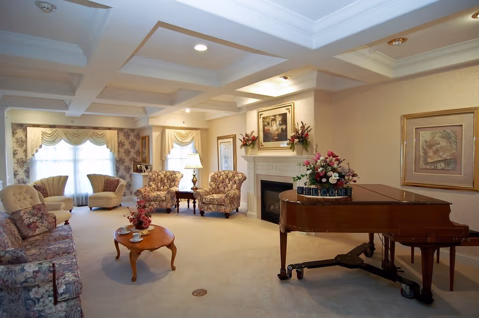 A cozy living room with floral upholstered chairs and sofa, a wooden coffee table with a flower arrangement and teacups, a grand piano with a floral centerpiece and a welcome sign, a fireplace with framed artwork and flower vases on the mantel, and large windows with draped curtains allowing natural light to fill the room.