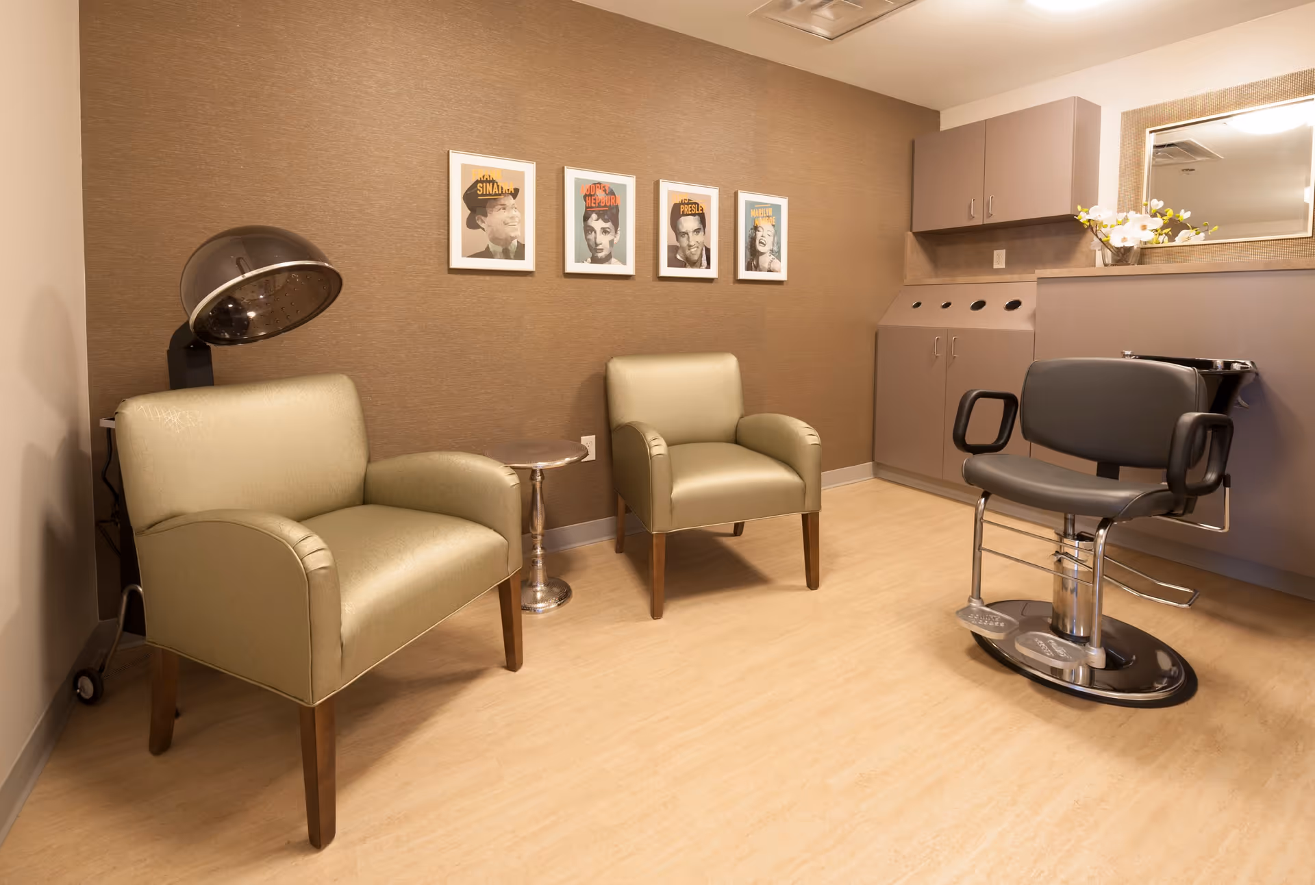 A salon room with two beige armchairs, a black salon chair, a hair dryer, a small round side table, and a cabinet with a mirror and flowers on top. Four framed portraits of Frank Sinatra, Audrey Hepburn, Elvis Presley, and Marilyn Monroe hang on the brown wall.