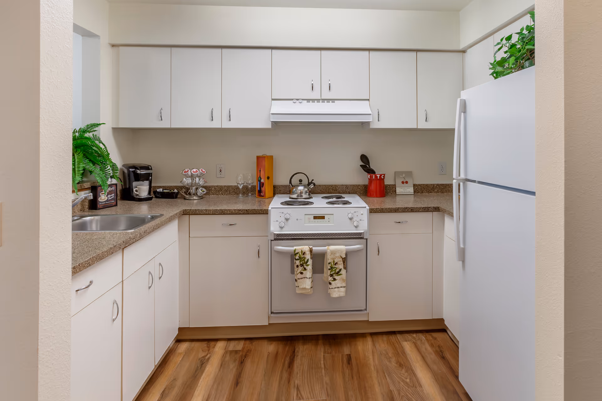 A clean and modern kitchen with white cabinets and drawers, a white stove with two decorative towels hanging on the oven handle, a white refrigerator, and a countertop with a coffee maker, a plant, wine glasses, and kitchen utensils. The floor is wood with a natural finish.