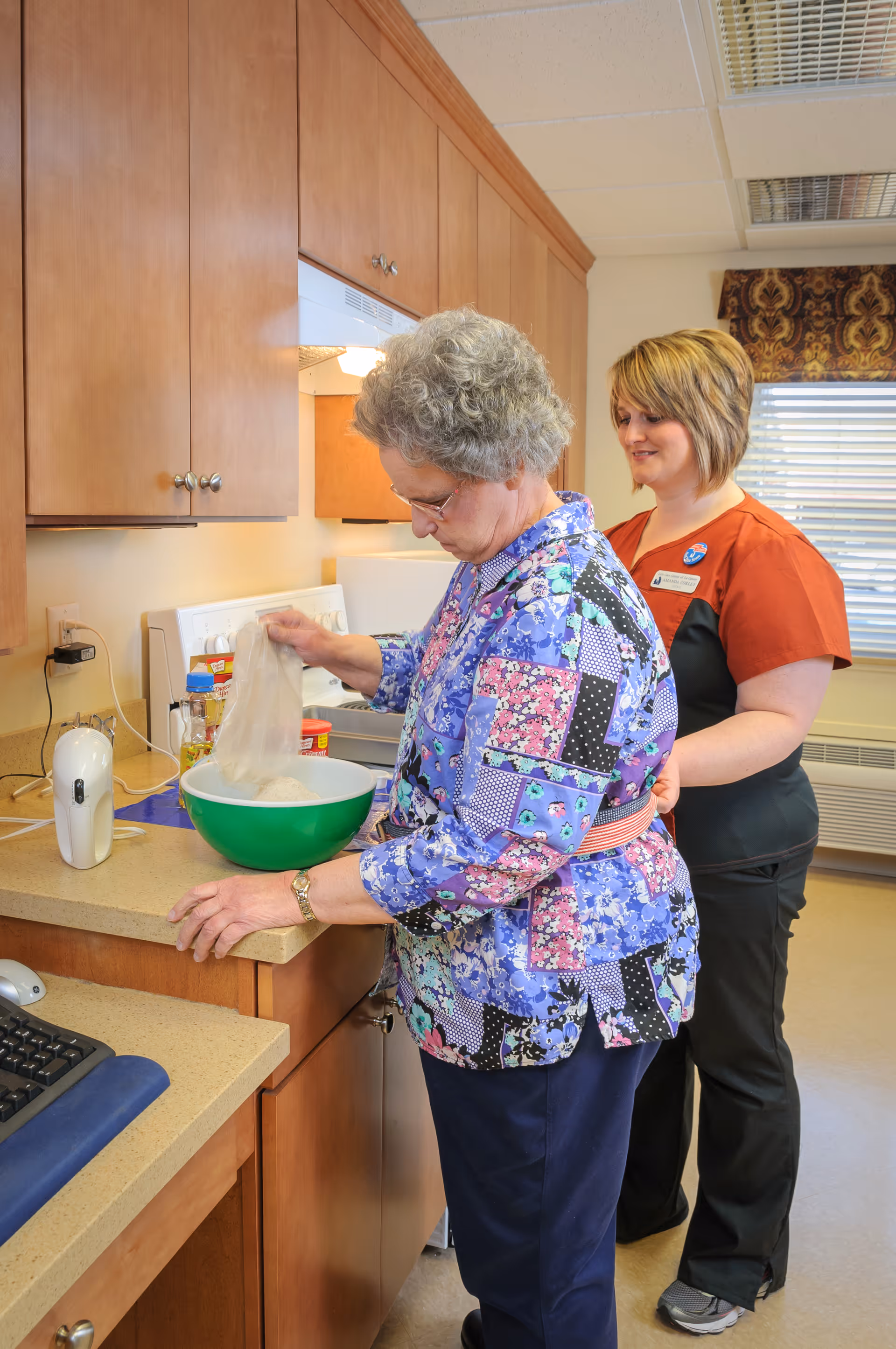 An elderly woman is standing at a kitchen counter mixing ingredients in a green bowl while a caregiver in an orange and black uniform stands behind her, smiling. The kitchen has wooden cabinets, a white stove, and a window with blinds and a patterned valance.
