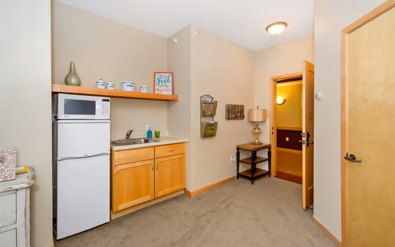 Small kitchenette area with a white microwave on top of a white mini refrigerator, a countertop with a sink, wooden cabinets below, and a wooden shelf above holding decorative jars and a framed sign. To the right, there is an open door leading to another room, a small table with a lamp, and wall-mounted organizers.
