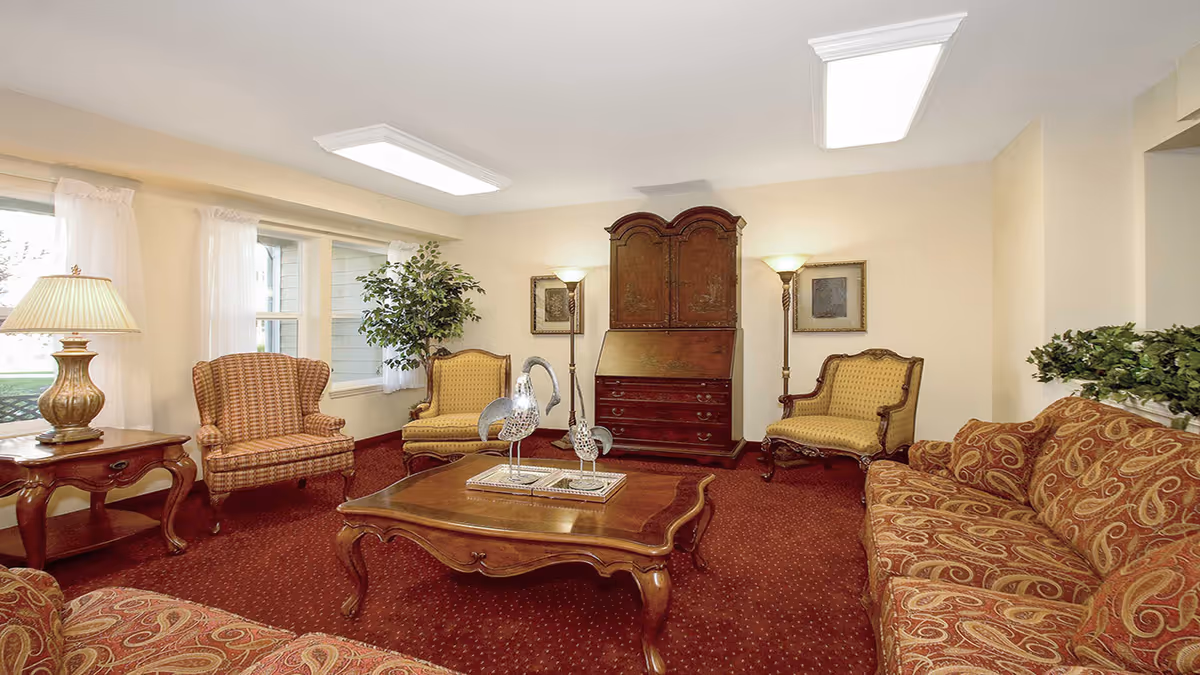 A cozy living room with patterned sofas and armchairs arranged around a wooden coffee table with decorative bird sculptures. The room has red carpet flooring, cream-colored walls, two floor lamps, framed artwork, a wooden cabinet, and a side table with a lamp. Large windows with sheer white curtains allow natural light to enter.