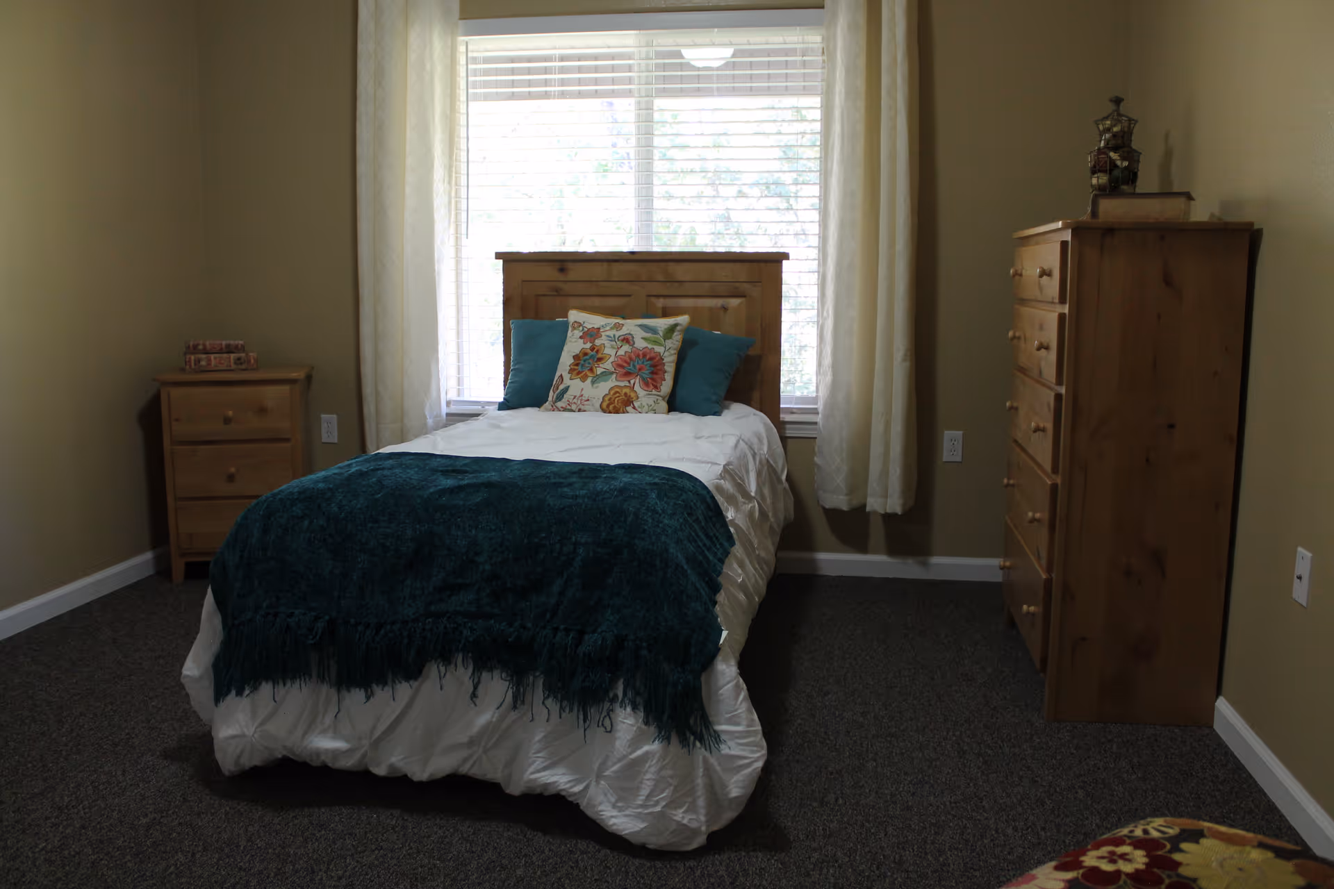 A small bedroom with a single wooden bed centered against a window with white curtains. The bed is made with white bedding, a dark teal throw blanket, and three decorative pillows. To the left of the bed is a small wooden nightstand with two drawers and some items on top. To the right is a taller wooden dresser with multiple drawers and a decorative item on top. The walls are beige and the floor is carpeted in dark gray.