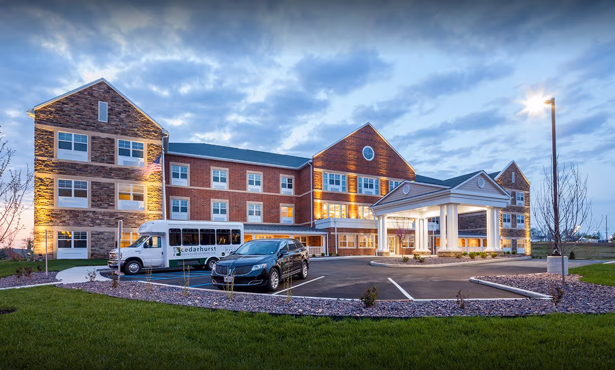 Exterior view of Cedarhurst Senior Living of St. Charles building at dusk, showing a three-story brick and stone facade with multiple windows, a covered entrance with white columns, a parking lot with a black car and a white shuttle bus, and a well-maintained lawn and landscaping.