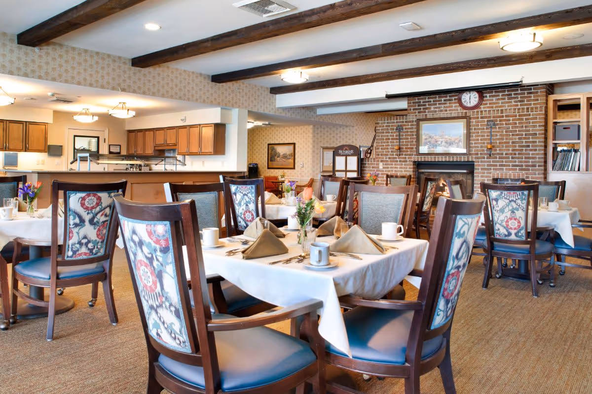Bright dining room with tables set for a meal, patterned wooden chairs, and a brick fireplace wall.