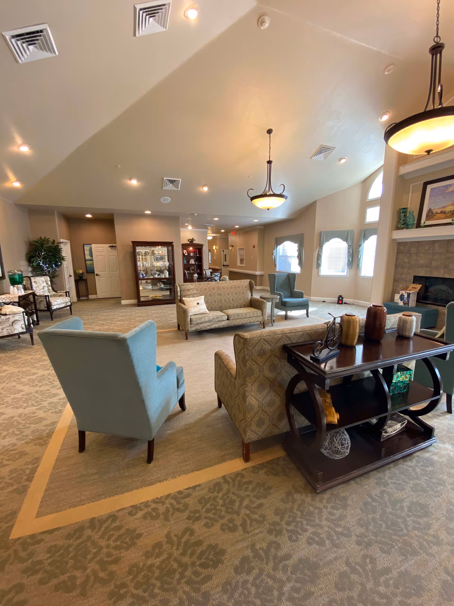 A spacious and well-lit living room area in a senior living facility with multiple upholstered chairs and sofas arranged around a dark wooden coffee table. The room features beige walls, patterned carpet, large windows with blue valances, a fireplace with decorative items on the mantel, and ceiling lights including hanging pendant lamps.