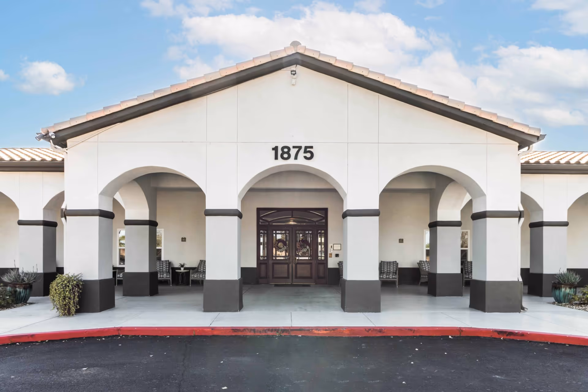 Front exterior view of a building with white walls and dark trim, featuring three large arches and the number 1875 displayed above the central arch. There are chairs and small tables under the arches, and the building has a tiled roof under a partly cloudy sky.
