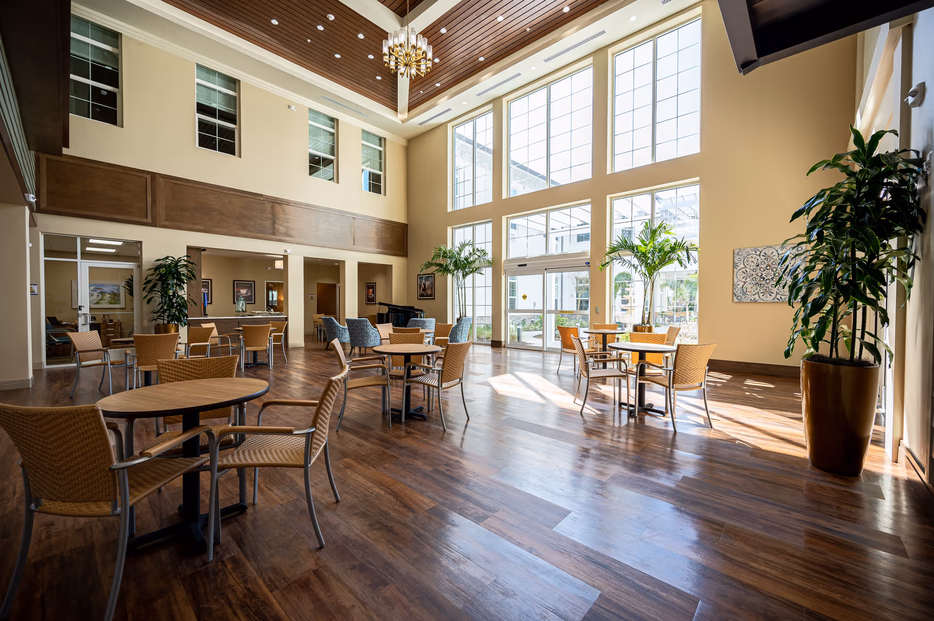 A spacious, well-lit common area with high ceilings and large windows allowing natural light to fill the room. The room features several round tables with wicker chairs, a few blue upholstered chairs, potted plants, and a grand piano in the background. The floor is wooden, and the walls are painted beige with wood paneling accents.