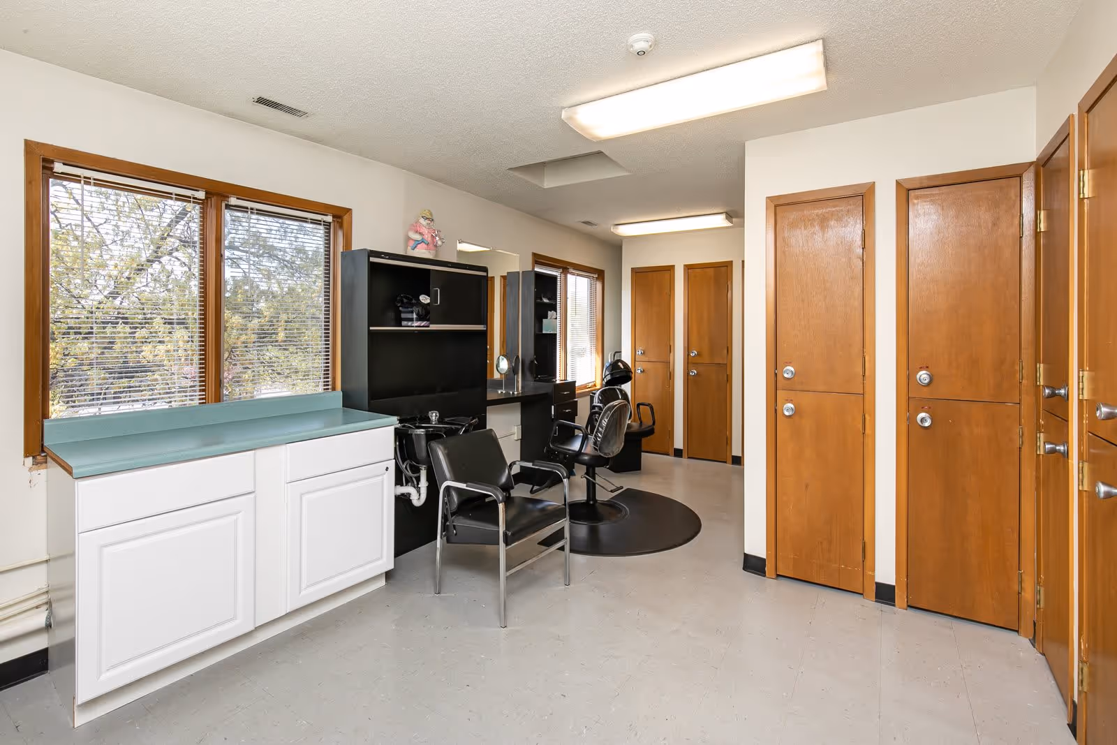 Interior view of a senior apartment salon area with two black salon chairs, a black hair washing station, a black shelving unit with a mirror, white cabinets with a green countertop, and several wooden doors. Two windows with blinds allow natural light into the room.
