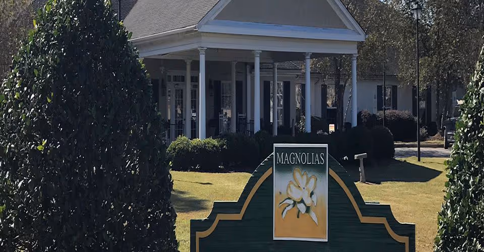 Exterior view of a senior living facility named Magnolias of Santee, showing a green and yellow sign with a magnolia flower in front of a building with white columns and a porch, surrounded by trimmed bushes and trees.