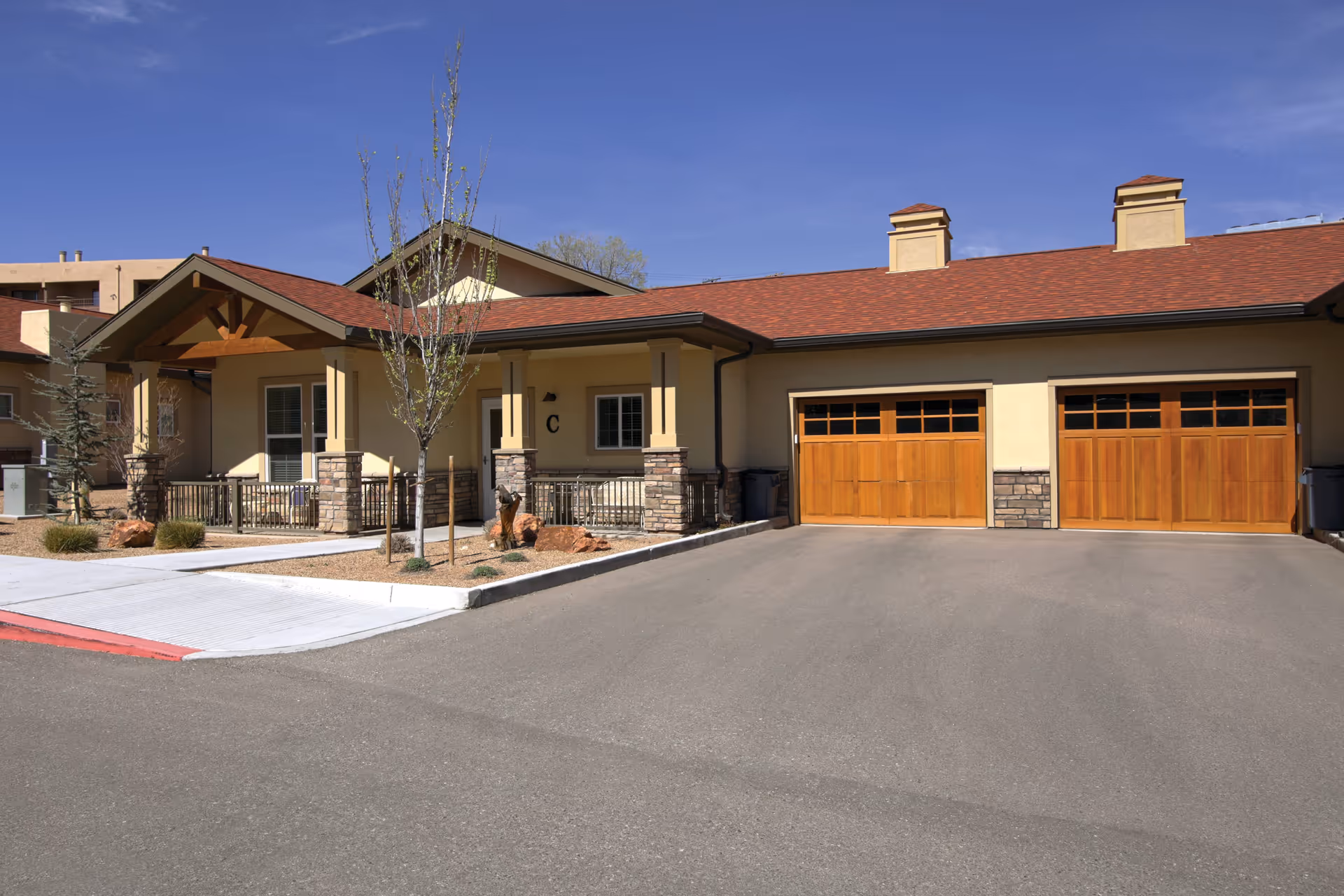 Exterior view of a single-story senior living facility building with a covered porch, two wooden garage doors, small trees, and a clear blue sky.