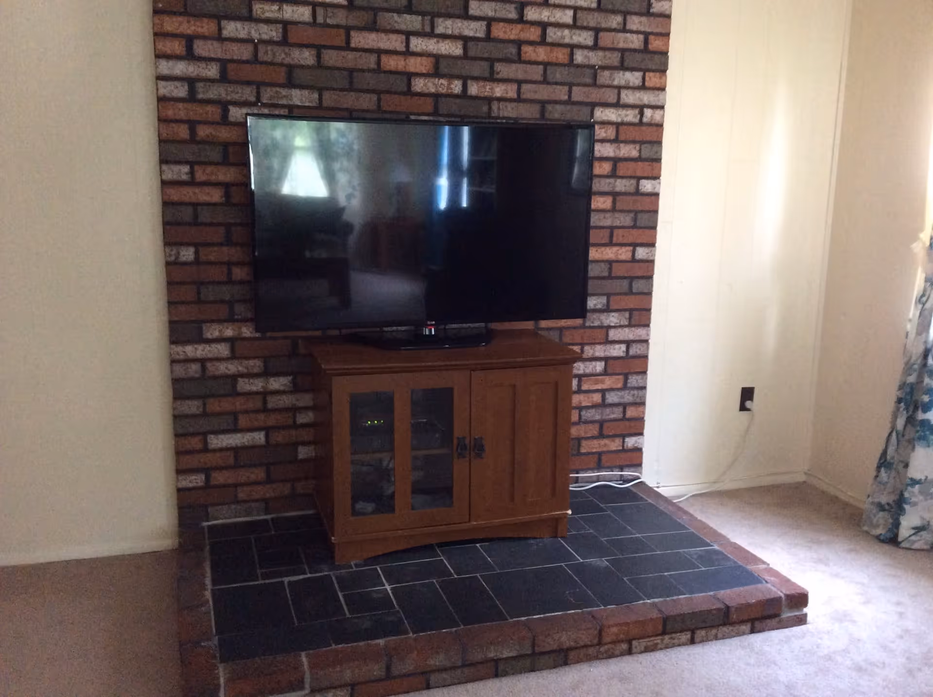 A flat-screen TV mounted on a brick wall above a wooden cabinet with glass doors, placed on a raised brick and tile platform in a room with light-colored walls and carpeted floor. A floral curtain is partially visible on the right side.