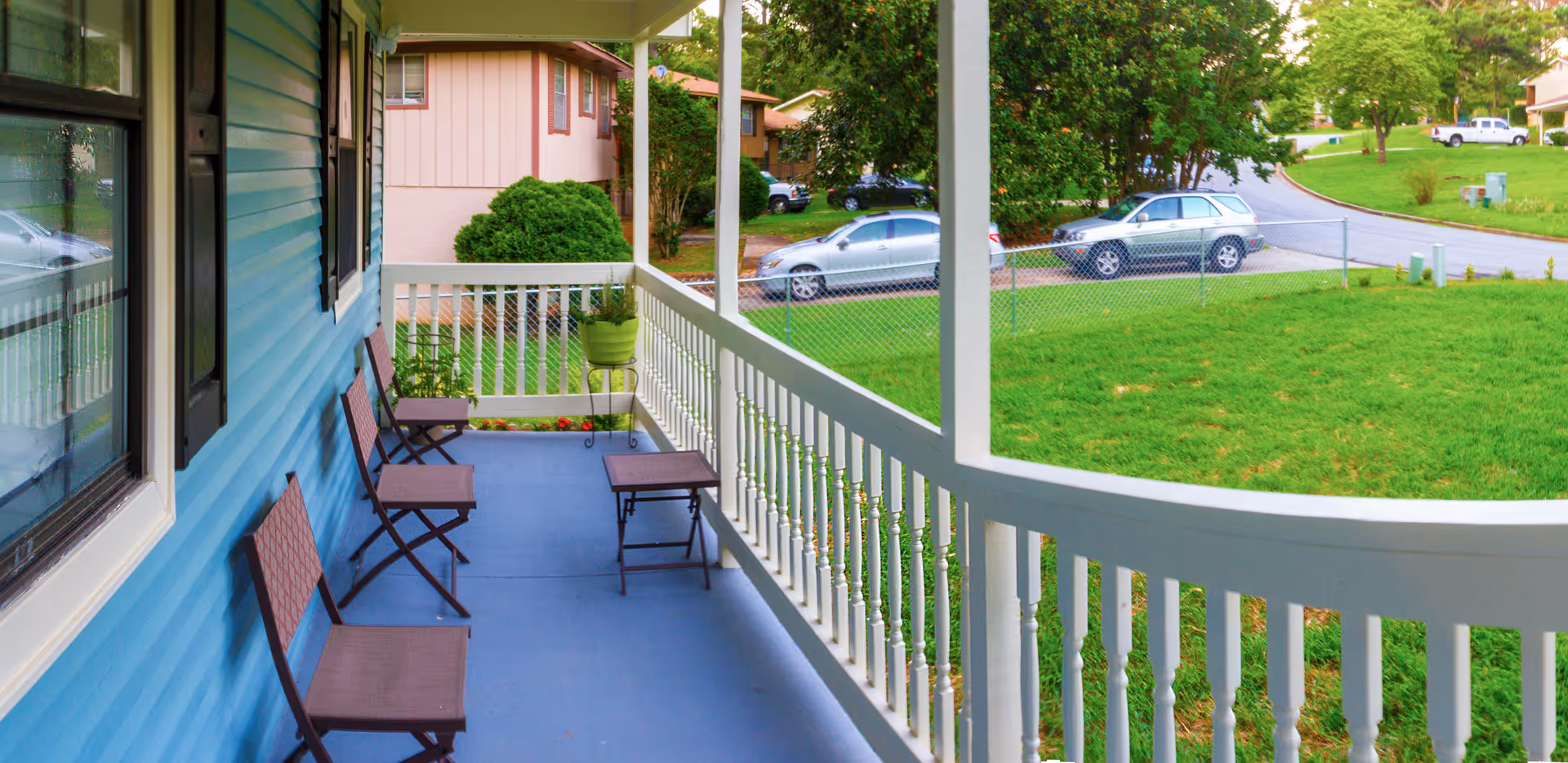 View of a covered porch with blue flooring and white railing, featuring four brown chairs and a small table. The porch overlooks a green lawn and a street with parked cars and trees in the background.