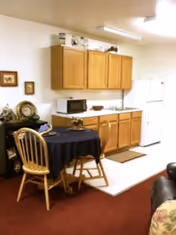 A small kitchen area with wooden cabinets, a microwave, a sink, and a white refrigerator. In front of the kitchen is a round table covered with a dark tablecloth and two wooden chairs. The floor is carpeted in a reddish color, and part of a couch with a floral cushion is visible on the right side.