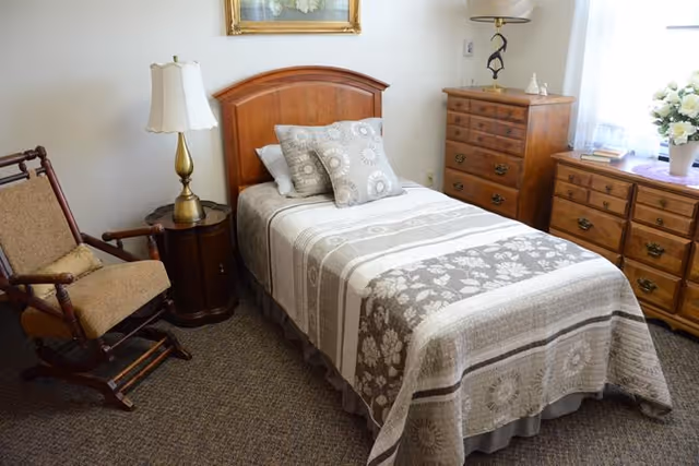 A cozy bedroom with a single bed covered in a patterned quilt and two decorative pillows. Next to the bed is a wooden nightstand with a brass lamp. There is a wooden rocking chair with a cushion on the left side. On the right side, there are two wooden dressers, one with a lamp and decorative items, and the other with a vase of white flowers near a window with sheer curtains. A framed painting hangs on the wall above the bed.