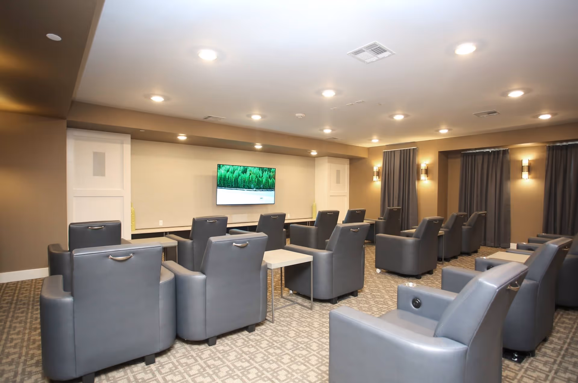 A modern lounge area with multiple gray leather armchairs arranged in rows facing a wall-mounted flat-screen TV displaying a nature scene. The room has beige walls, recessed ceiling lights, wall sconces, and windows covered with dark curtains. Small tables are placed between some chairs.