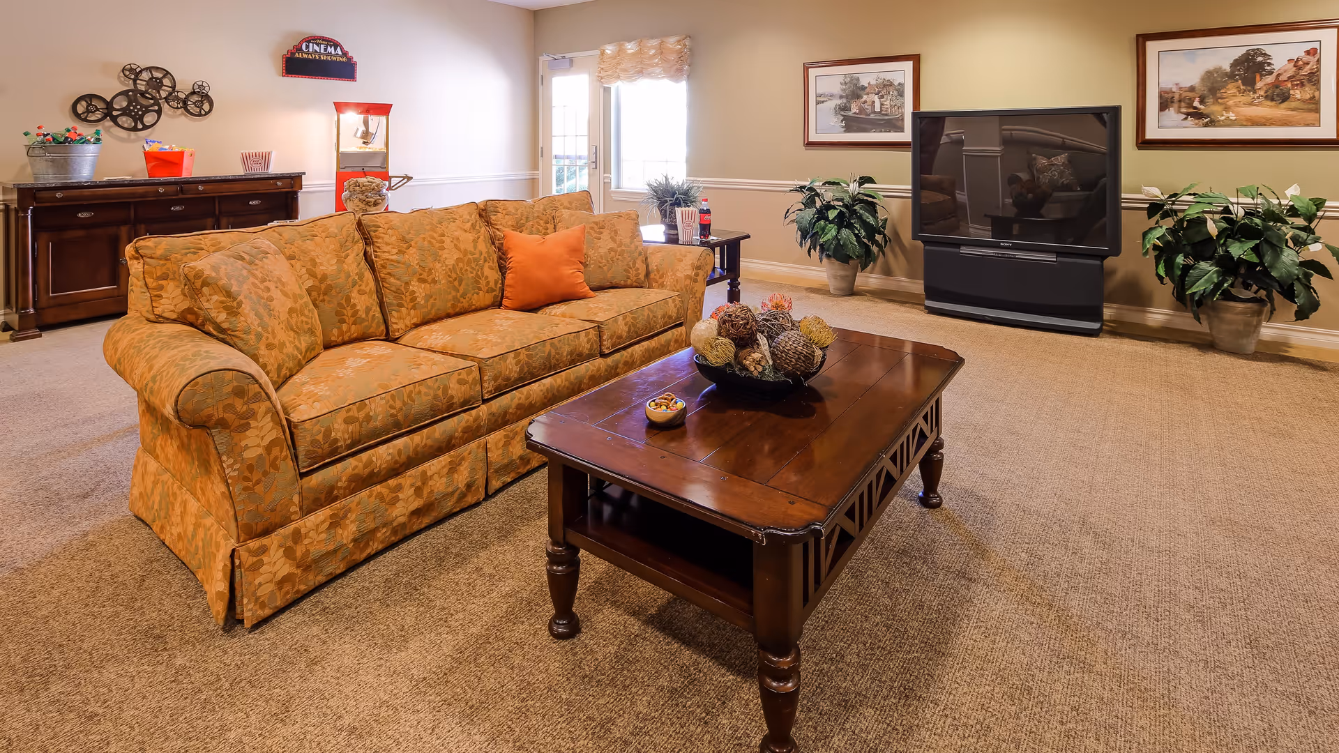 A cozy living room with a patterned orange sofa, a wooden coffee table with decorative items, a large flat-screen TV on a stand, two potted plants, framed artwork on the walls, and a sideboard with a popcorn machine and decorative items.