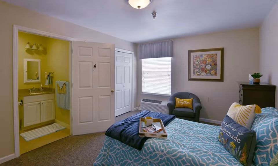 A cozy bedroom in an assisted living facility featuring a bed with blue and white patterned bedding and decorative pillows, a tray with a cup and glasses on the bed, a comfortable armchair with a yellow pillow, a framed floral artwork on the wall, a window with blinds and a valance, a dark wooden dresser with a small plant, and an open door leading to a bathroom with a sink, mirror, and towels.