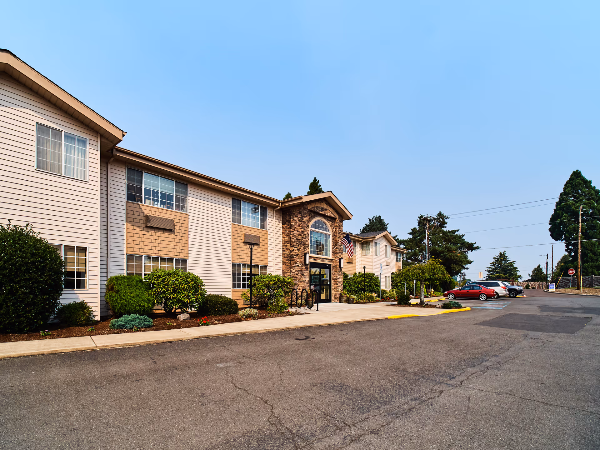 Exterior view of a two-story senior living facility building with beige siding and stone accents around the main entrance. There are several windows, some bushes and small trees planted along the front, a paved driveway, and a few parked cars. The sky is clear and blue.
