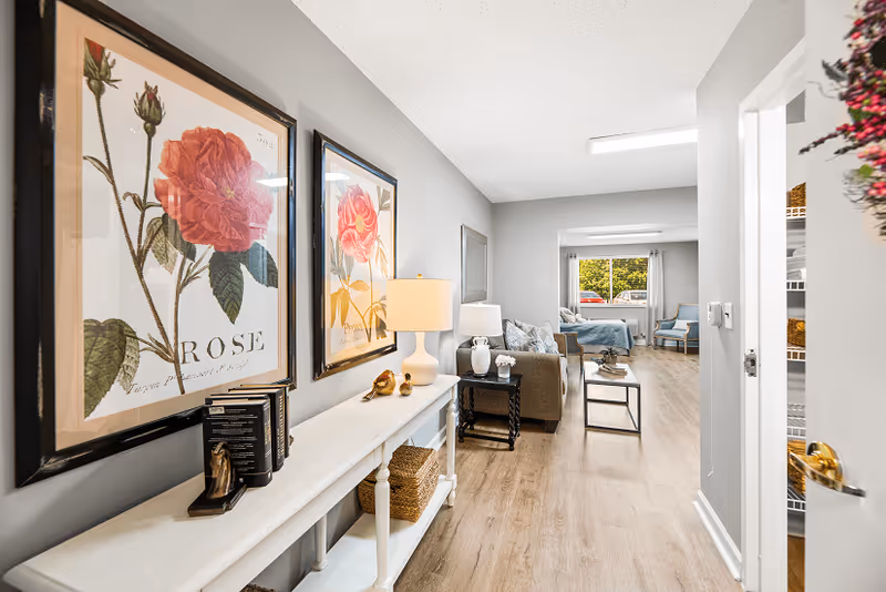 View down a hallway in a senior living facility apartment featuring light wood flooring, gray walls, and floral artwork on the left wall. A white console table with decorative items and a lamp is positioned under the artwork. Further down, a living area with a sofa, side table, coffee table, and a bed near a window with curtains is visible. The right side shows an open door leading to a pantry or closet with shelves and baskets.