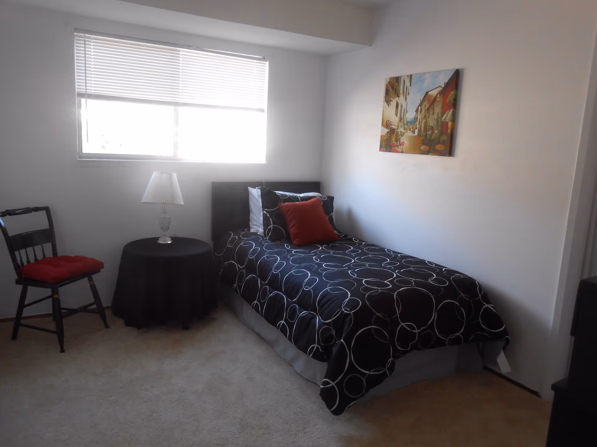 A small bedroom with a single bed covered in a black comforter with white circular patterns and a red pillow. Next to the bed is a round table with a black tablecloth and a lamp on top. A wooden chair with a red cushion is placed beside the table. A window with blinds is on the left wall, and a colorful painting of a street scene hangs on the right wall.