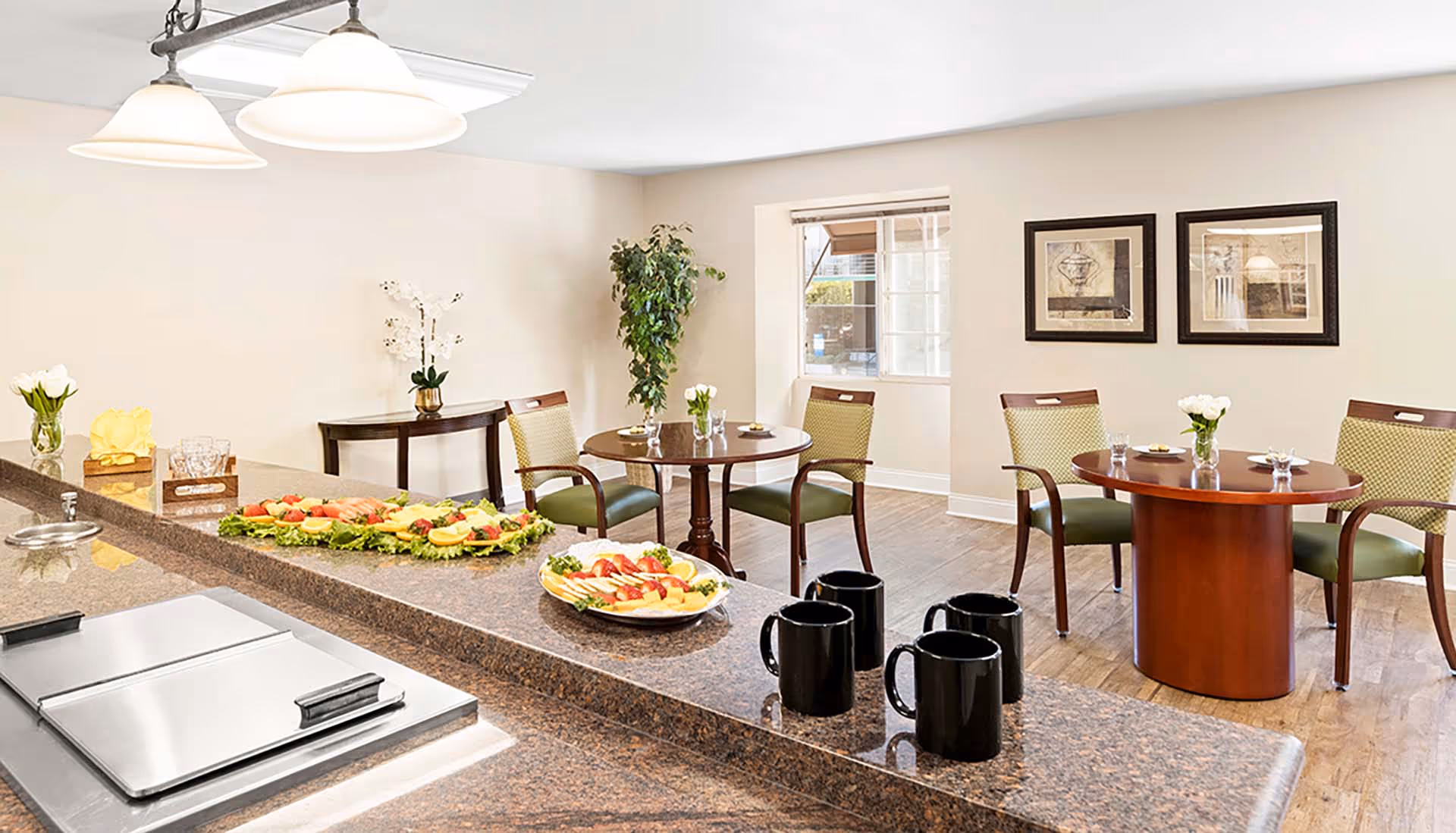 A bright dining area with a granite countertop in the foreground displaying plates of fresh fruit and four black mugs. In the background, there are two round wooden tables each with two green cushioned chairs, small flower vases, and framed artwork on the wall. A window lets in natural light, and a potted plant is placed near the corner.