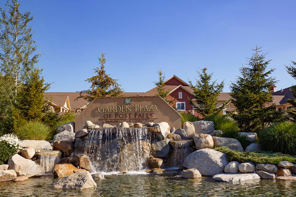 Outdoor view of the Garden Plaza of Post Falls senior living community sign with a decorative waterfall feature surrounded by rocks, greenery, and trees, with the facility building visible in the background under a clear blue sky.