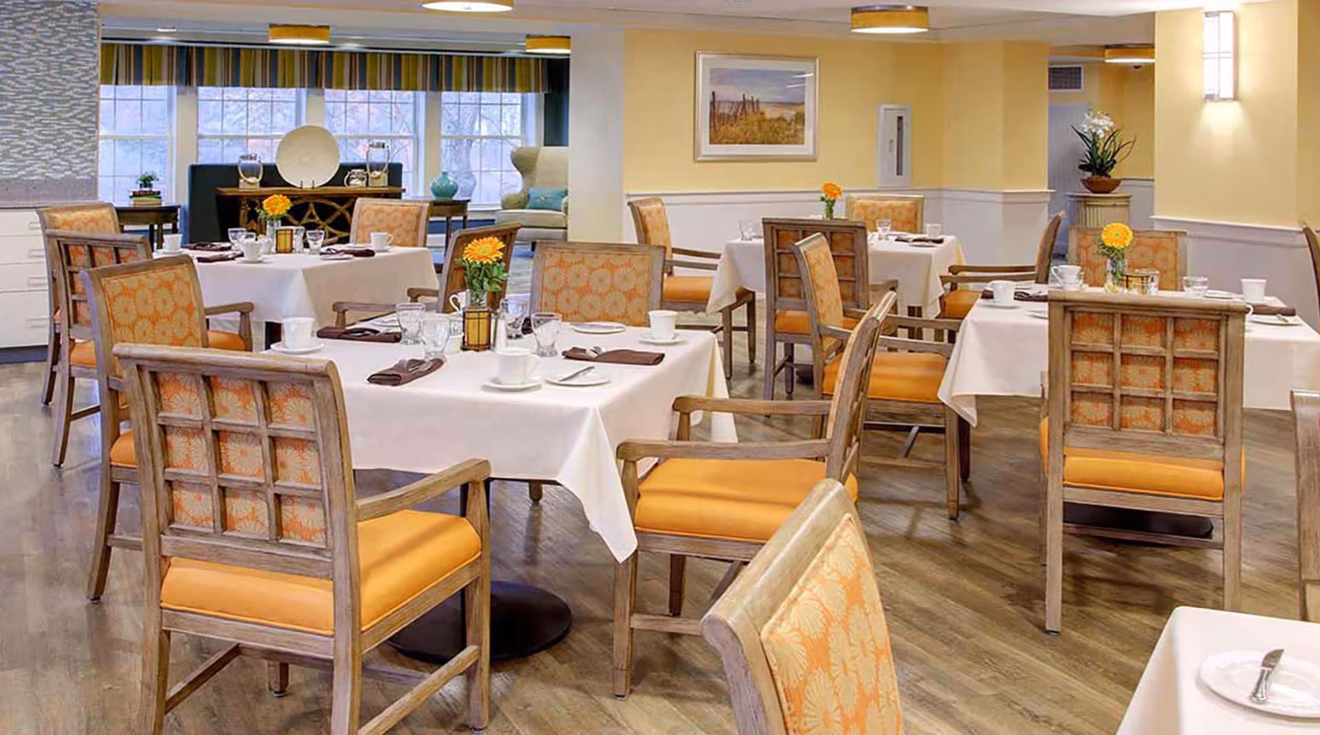 A bright and neatly arranged dining room with multiple tables covered in white tablecloths, each set with cups, glasses, plates, and napkins. The chairs have wooden frames with orange cushioned seats and patterned backs. The room has large windows with striped valances, light yellow walls, and decorative plants and artwork.