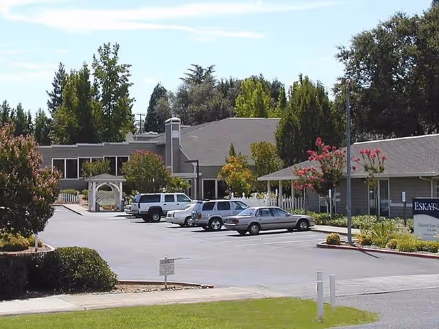 Exterior view of Eskaton Care Center Fair Oaks showing a parking lot with several parked cars, surrounded by trees and bushes, with the building and a sign visible on the right side.
