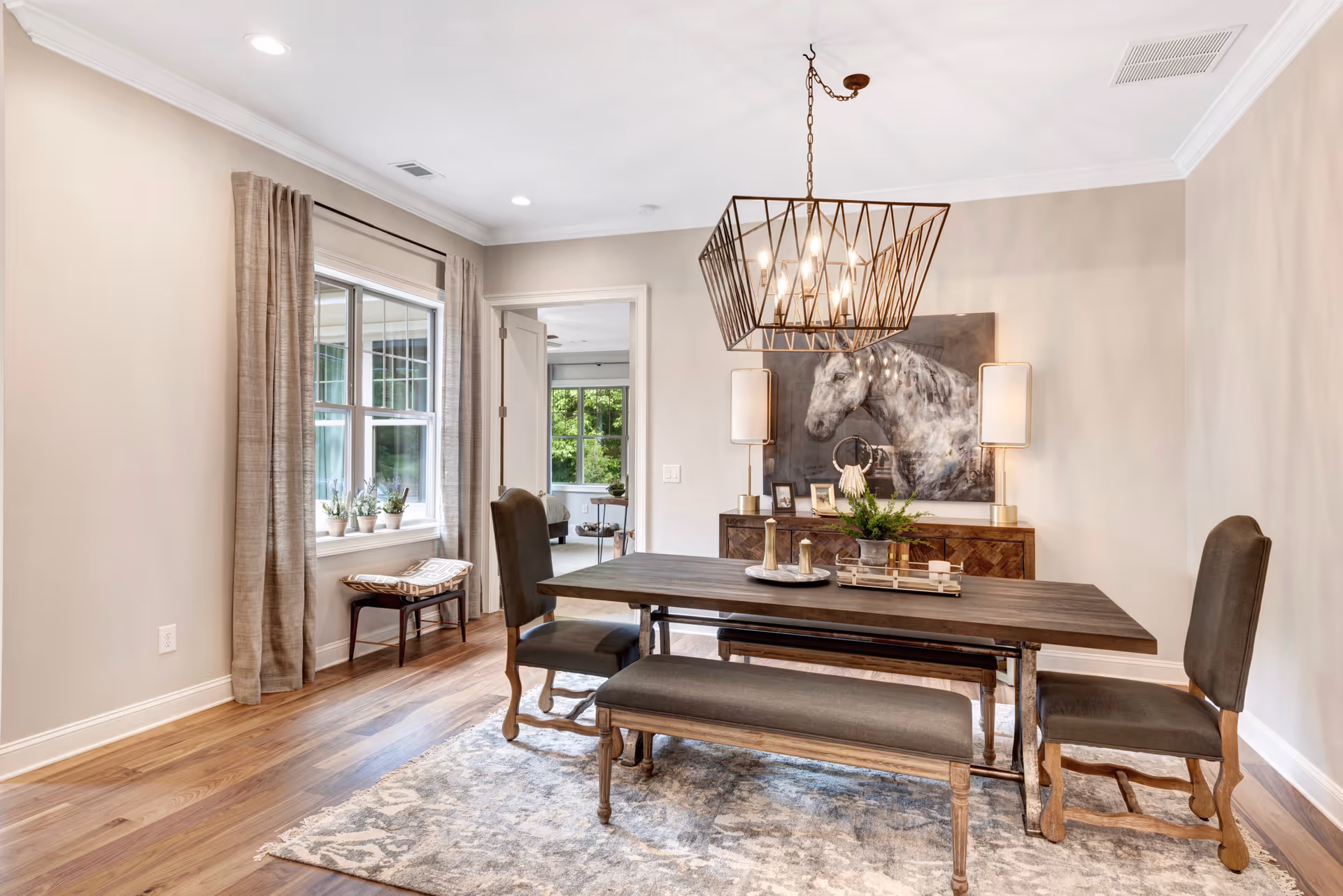 A bright dining room with a wooden dining table, two upholstered chairs, and a bench. The room features hardwood floors, a large window with beige curtains, a modern geometric chandelier, and a sideboard with two lamps and a large horse painting above it. There is a doorway leading to another room with a window showing greenery outside.