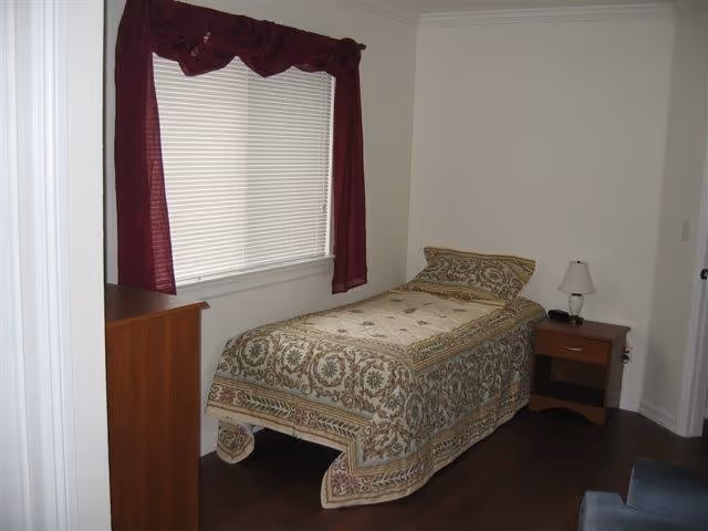 Small bedroom with a twin bed covered in a patterned bedspread, a nightstand with lamp, dresser, and a window with blinds and a maroon valance.