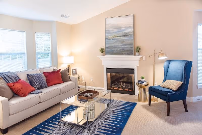A cozy living room with a beige sofa adorned with red and blue pillows, a blue patterned rug, a glass coffee table, a blue armchair with a beige pillow, a fireplace with a painting above it, and two windows letting in natural light.