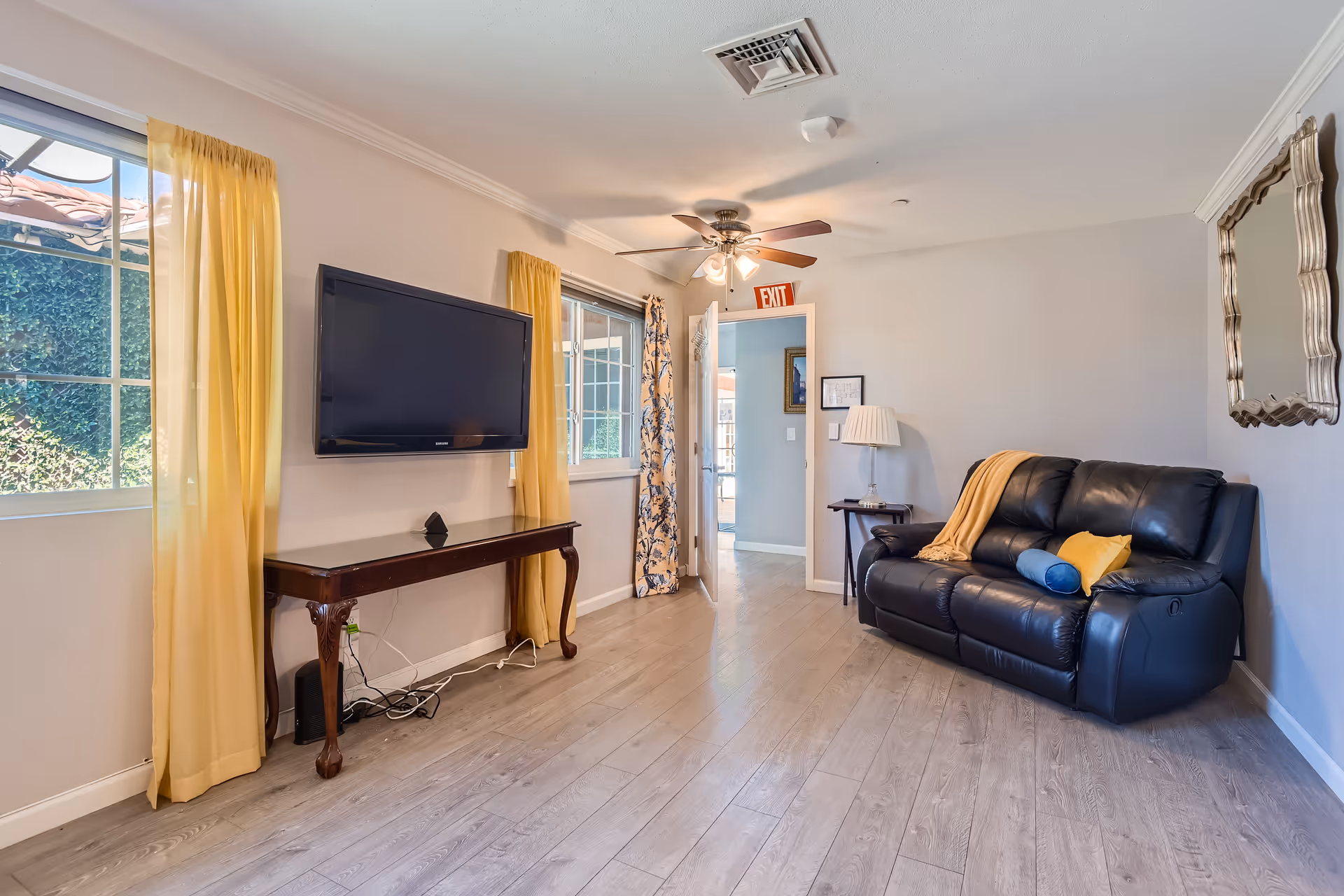 A cozy living room with light wood flooring, a black leather couch with yellow and blue pillows, a side table with a lamp, a wall-mounted flat screen TV above a wooden console table, and two windows with yellow curtains letting in natural light. A ceiling fan with lights is mounted on the ceiling, and an exit sign is visible above an open door leading to another room.