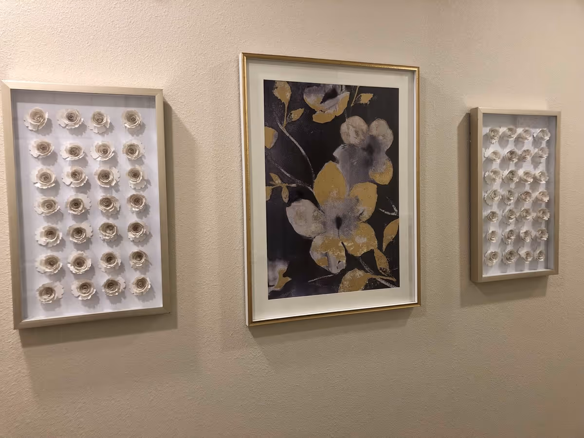 Three framed wall decorations hanging on a beige wall. The center frame contains a floral painting with large flowers in shades of yellow, gray, and black. The two side frames each contain a grid of white 3D flower sculptures arranged in rows.