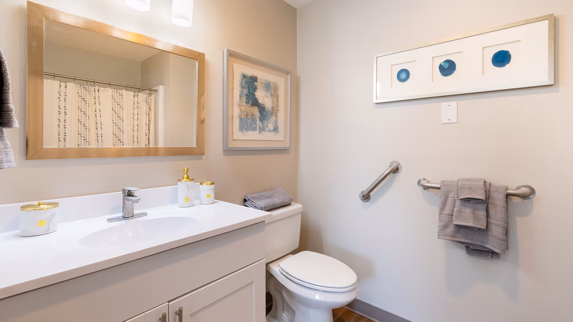 A clean and modern bathroom featuring a white vanity with a sink, a large rectangular mirror with a gold frame, a toilet with folded gray towels on top, a towel rack with gray towels, and two framed abstract artworks on the walls.