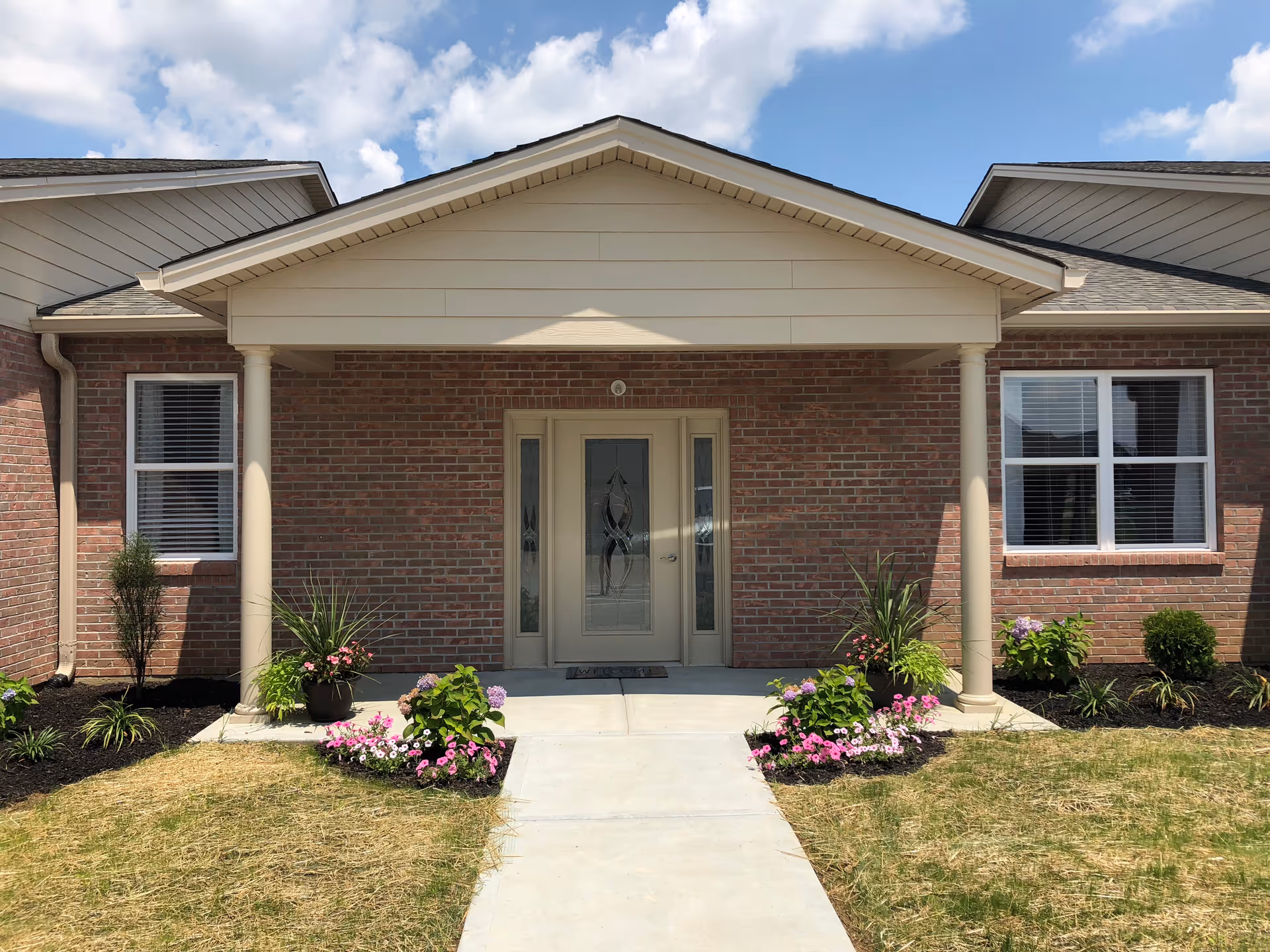 Covered front entrance of a brick senior living building with columns, a decorative glass door, and flower beds.