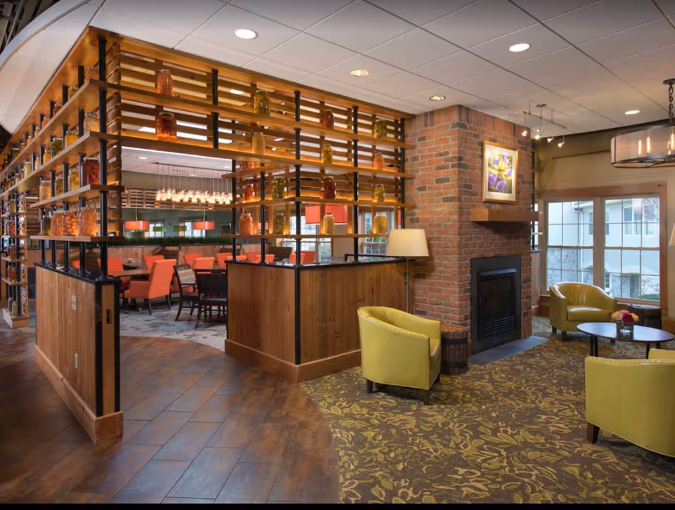 Interior view of a senior living facility lounge area with green armchairs around a small round table on a patterned carpet near a brick fireplace. Adjacent to the lounge is a dining area with orange chairs and a wooden partition decorated with jars of preserved vegetables.